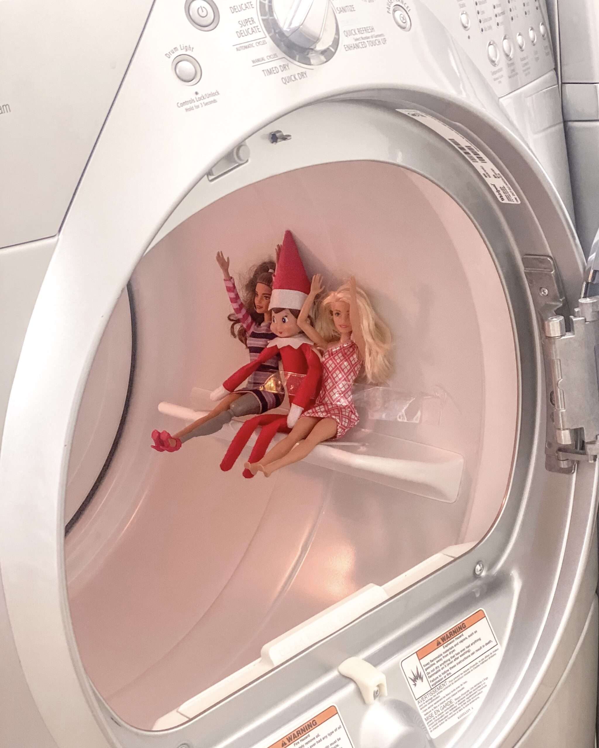 Turn your laundry room into an amusement park for your elf. Fun & easy Elf on the Shelf ideas for your toddler or little kid. Elf swinging on a roll of toilet paper. Quick to setup and lots of naughty and nice ideas, perfect to bring some Christmas magic to your home. #ElfOnTheShelf #ElfIdeas #ElfOnTheShelfIdeas #Christmas #ChristmasMagic