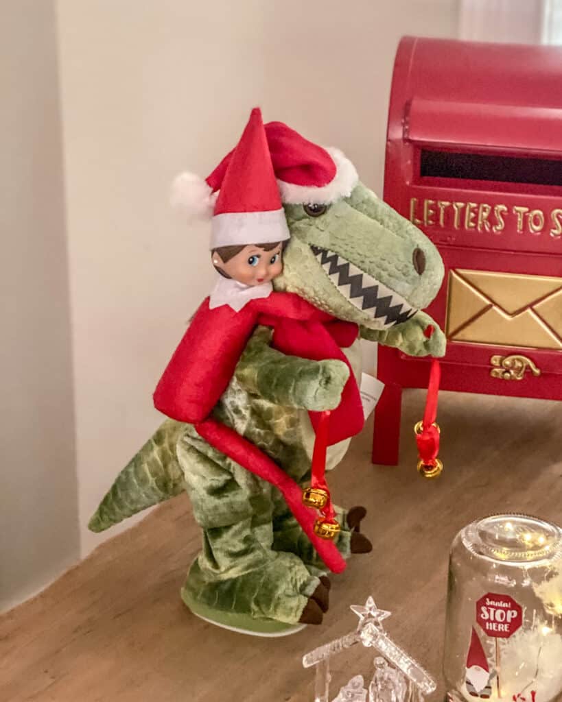 Elf riding on a dinosaur. Fun & easy Elf on the Shelf ideas for your toddler or little kid. Quick to setup and lots of naughty and nice ideas, perfect to bring some Christmas magic to your home. #ElfOnTheShelf #ElfIdeas #ElfOnTheShelfIdeas #Christmas #ChristmasMagic