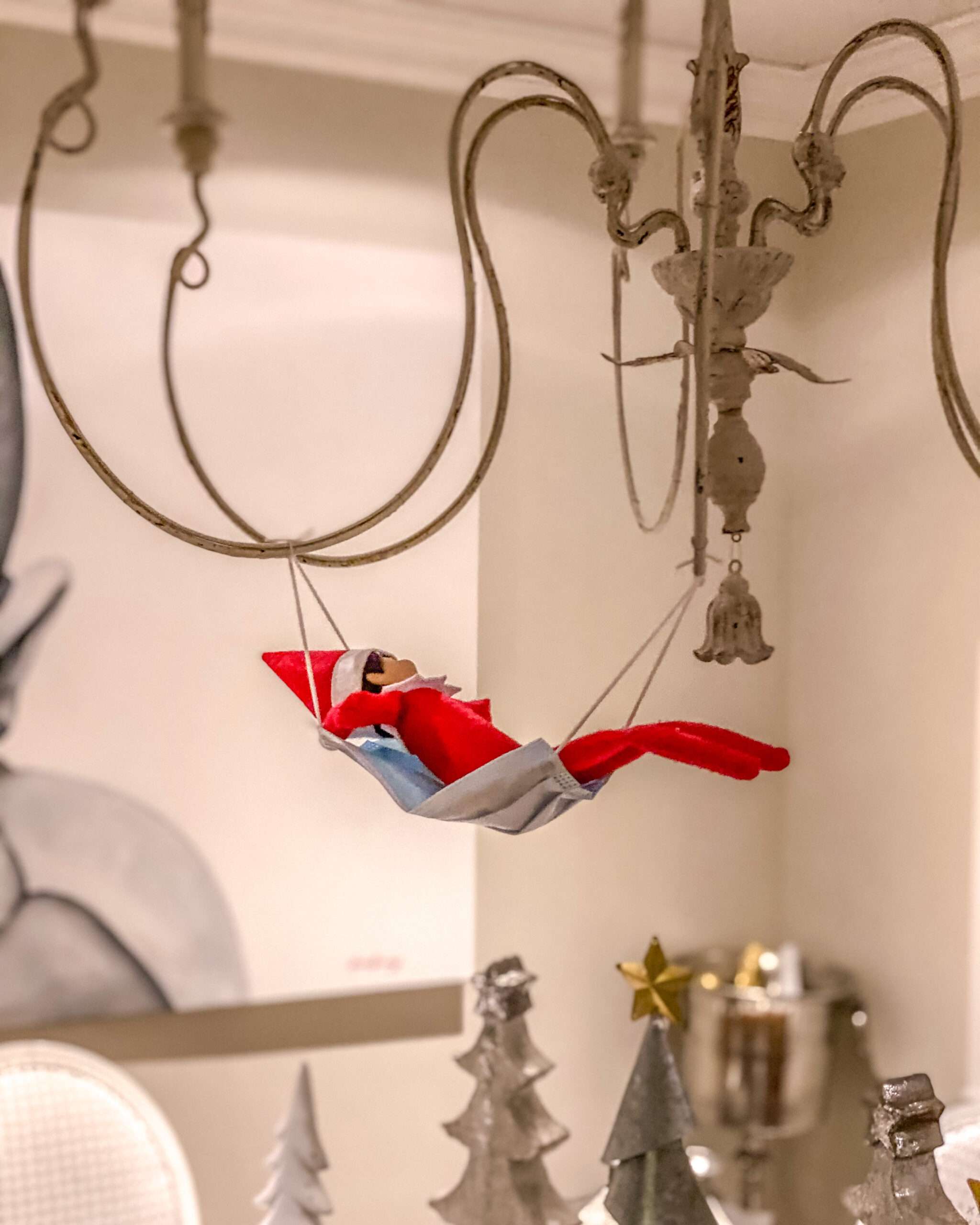 Disposable face mask hanging on a chandelier as a elf hammock. Fun & easy Elf on the Shelf ideas for your toddler or little kid. Quick to setup and lots of naughty and nice ideas, perfect to bring some Christmas magic to your home. #ElfOnTheShelf #ElfIdeas #ElfOnTheShelfIdeas #Christmas #ChristmasMagic