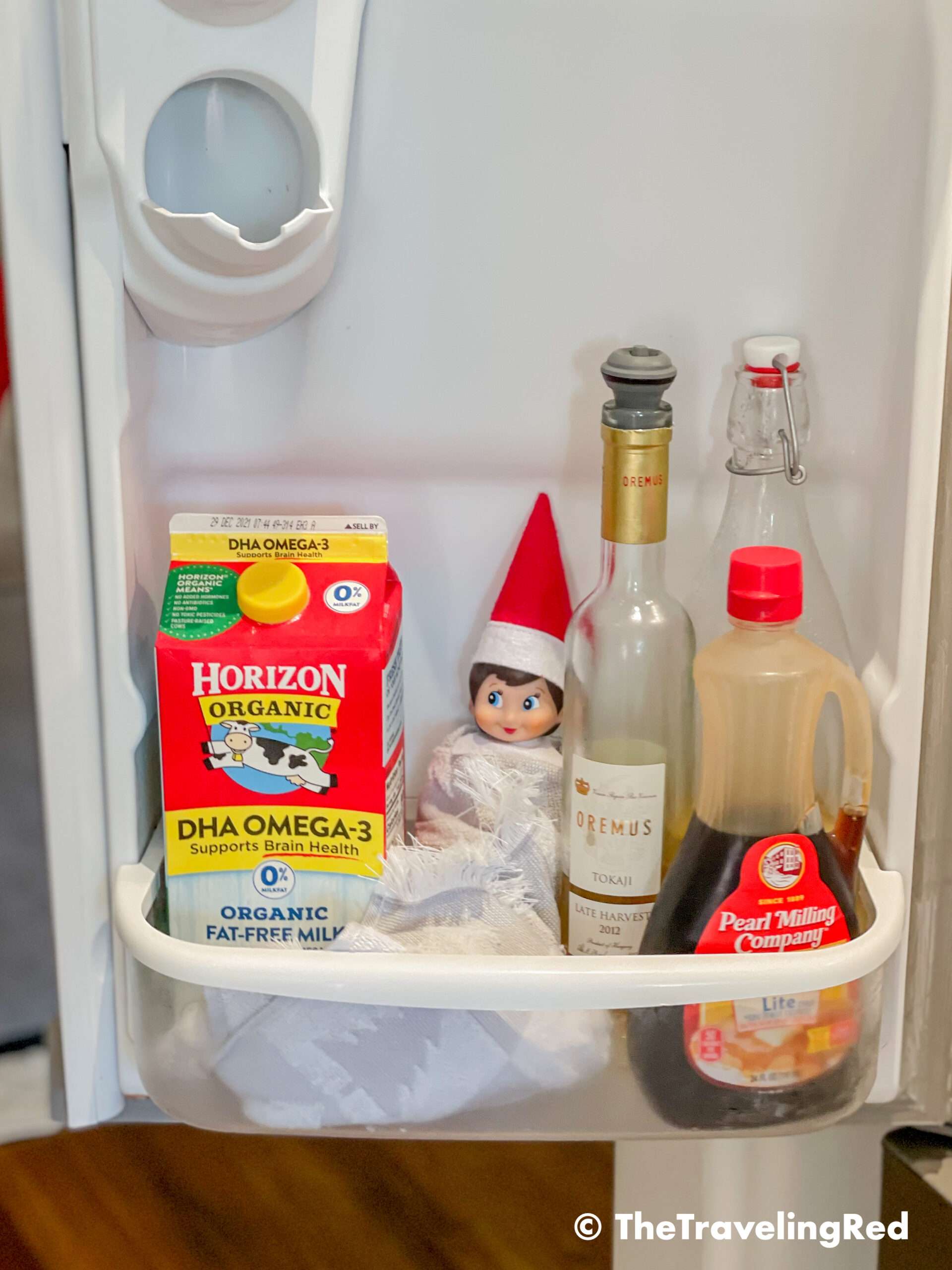 Naughty Elf on the shelf hiding in the fridge using a kitchen hand towel as a blanket. Fun and easy elf on the shelf ideas for a naughty elf that are quick and easy using things you have at home.