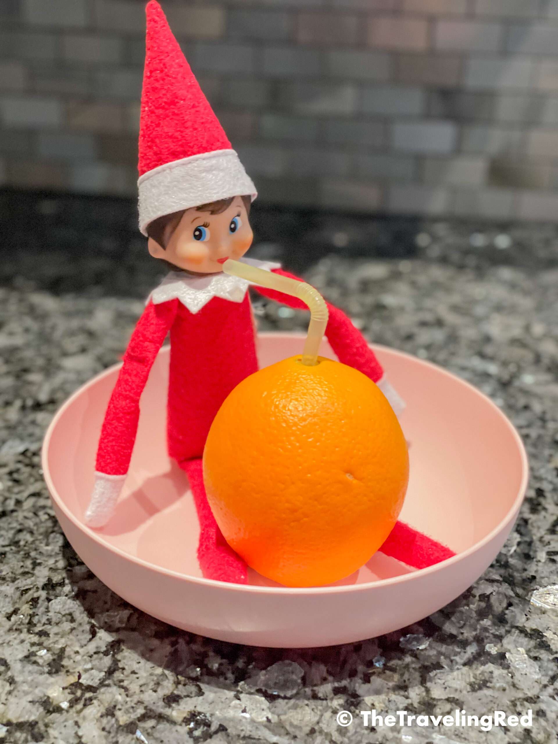 Naughty Elf on the shelf drinking orange juice right out of an orange by inserting a straw. Fun and easy elf on the shelf ideas for a naughty elf that are quick and easy using things you have at home.