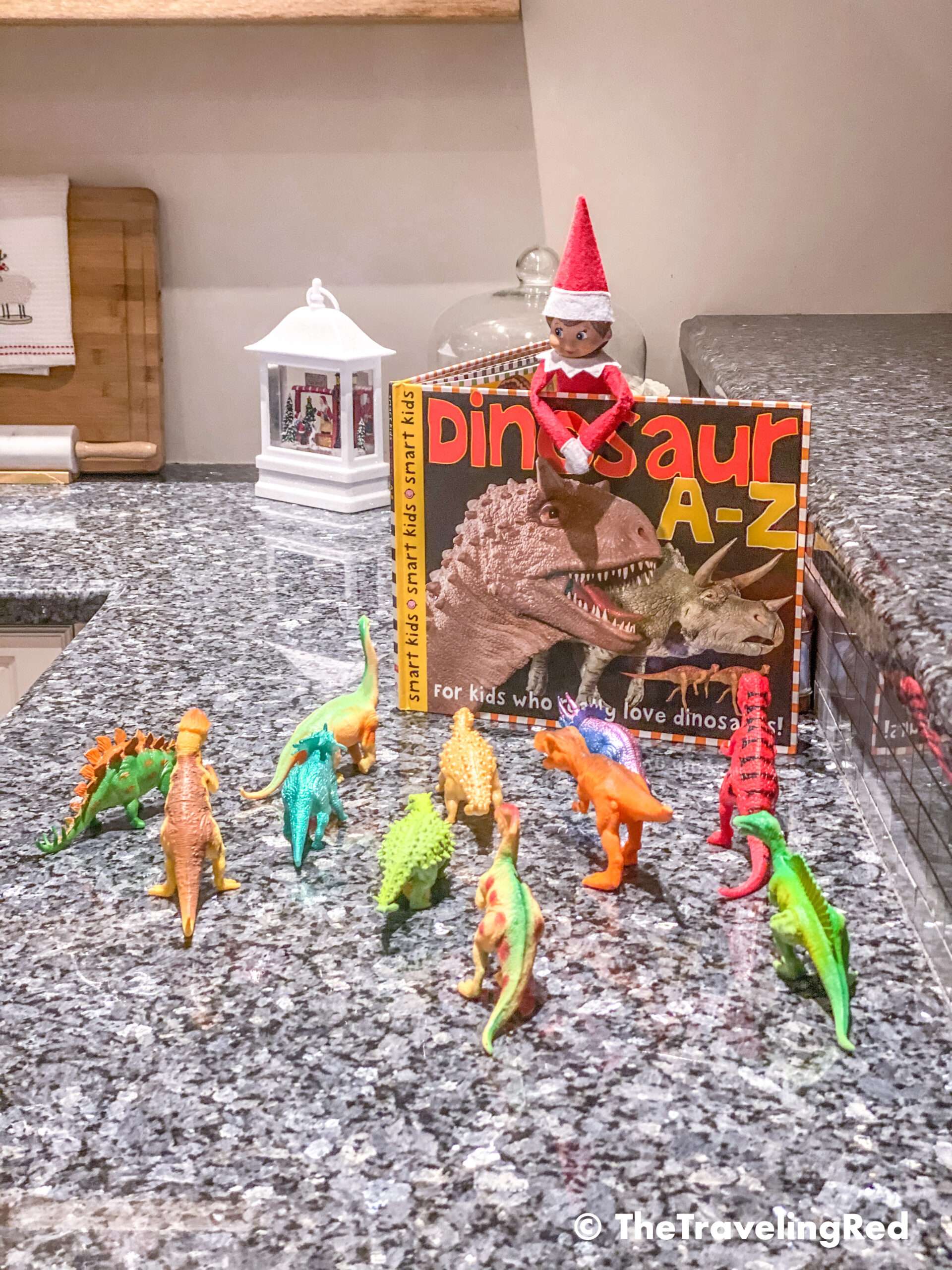 Naughty Elf on the shelf reading a dinosaur book to the dinosaurs. Fun and easy elf on the shelf ideas for a naughty elf that are quick and easy using things you have at home.
